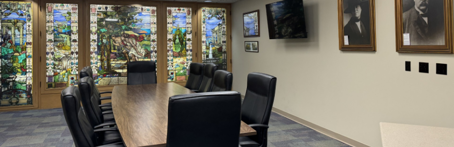 Board room with large wooden table and ten chair surrounding it and television on wall.