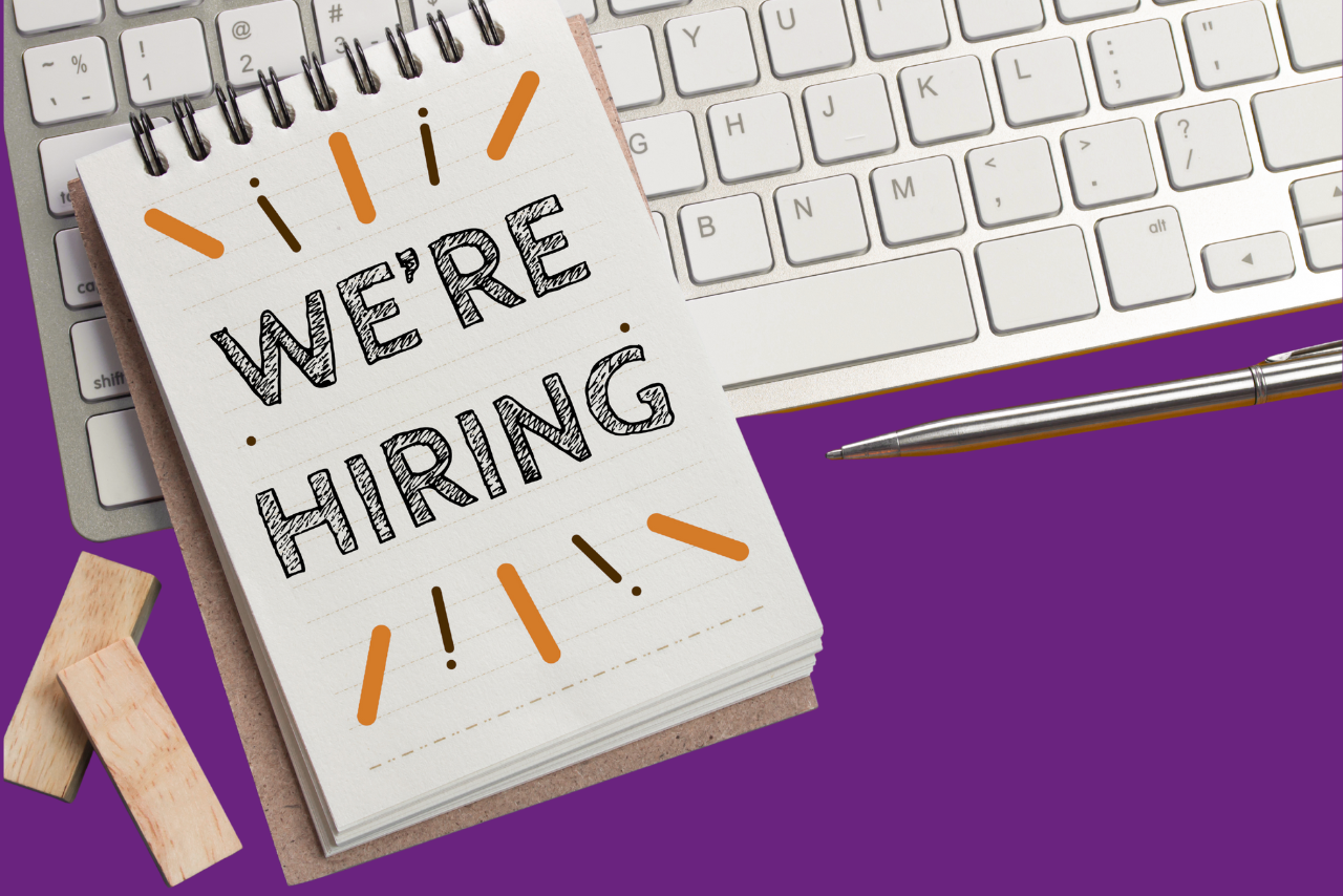 A notepad that has 'We're Hiring' written on it laying on a computer keyboard.